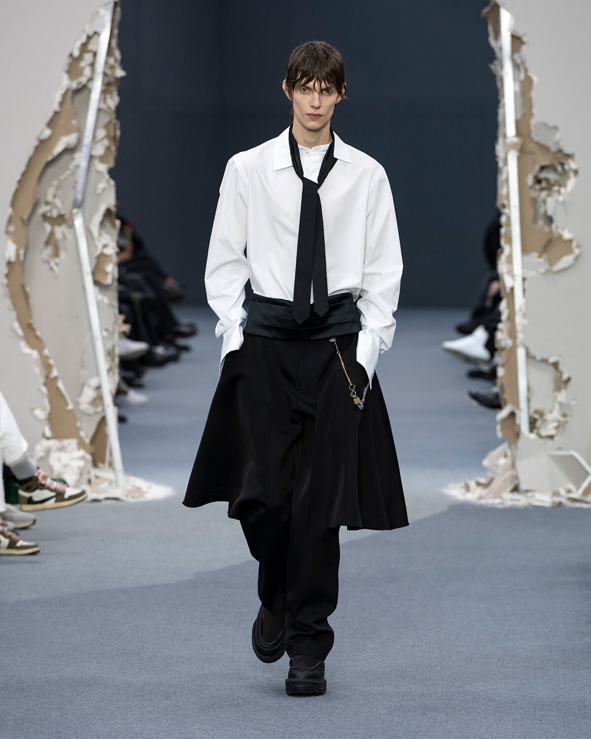 Male model on runway wearing a white dress shirt, black tie, and voluminous black trousers with a chain accessory