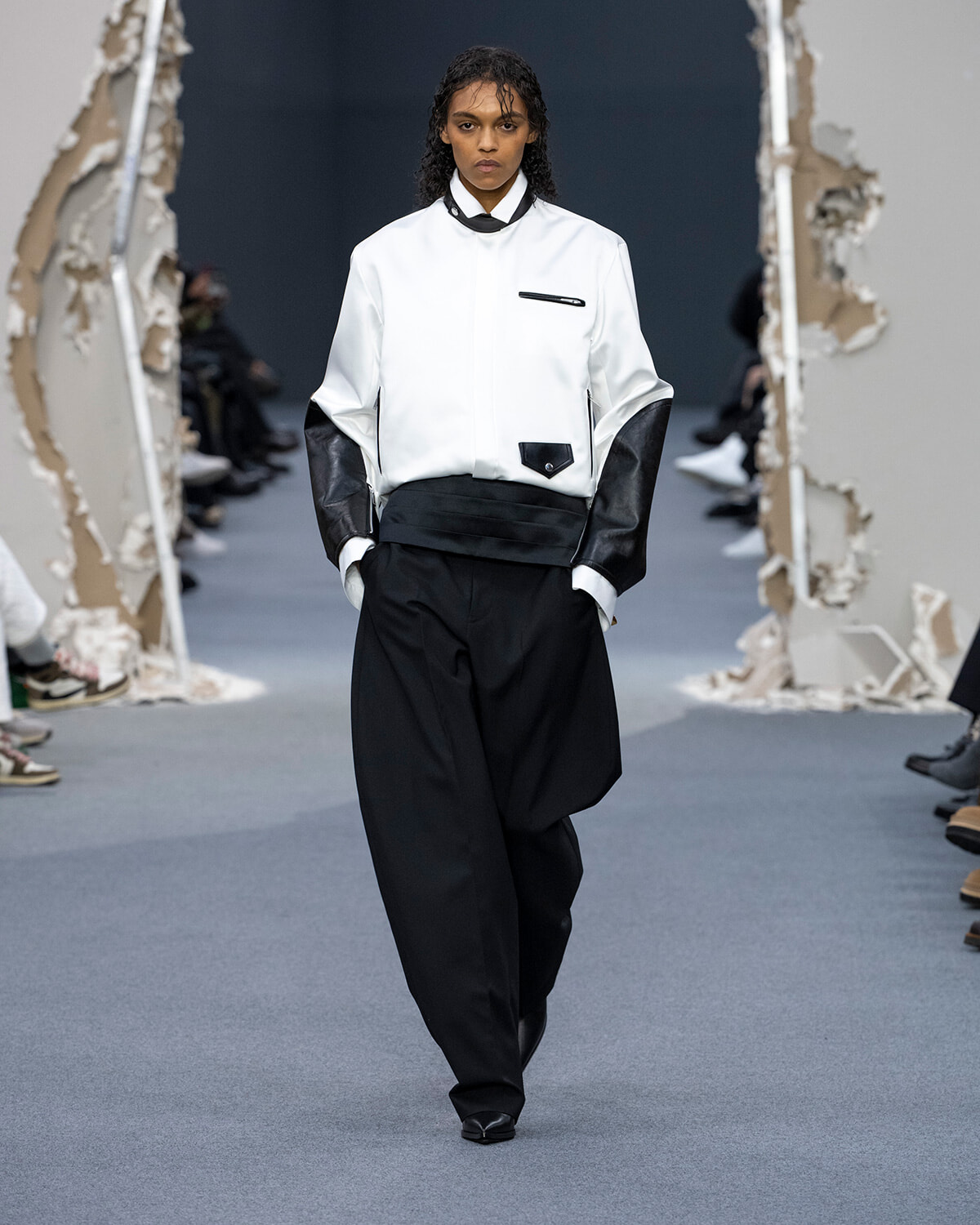 Model walks runway in a white structured jacket with black accents and oversized black pants, minimalist monochrome look.