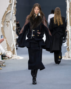 Model walks a runway wearing a navy coat with brown leather panels, fur collar, gold buttons, and tall black boots.