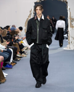 Male model in a black utility jacket and baggy pants walks a runway during a fashion show, with spectators seated along the sides.