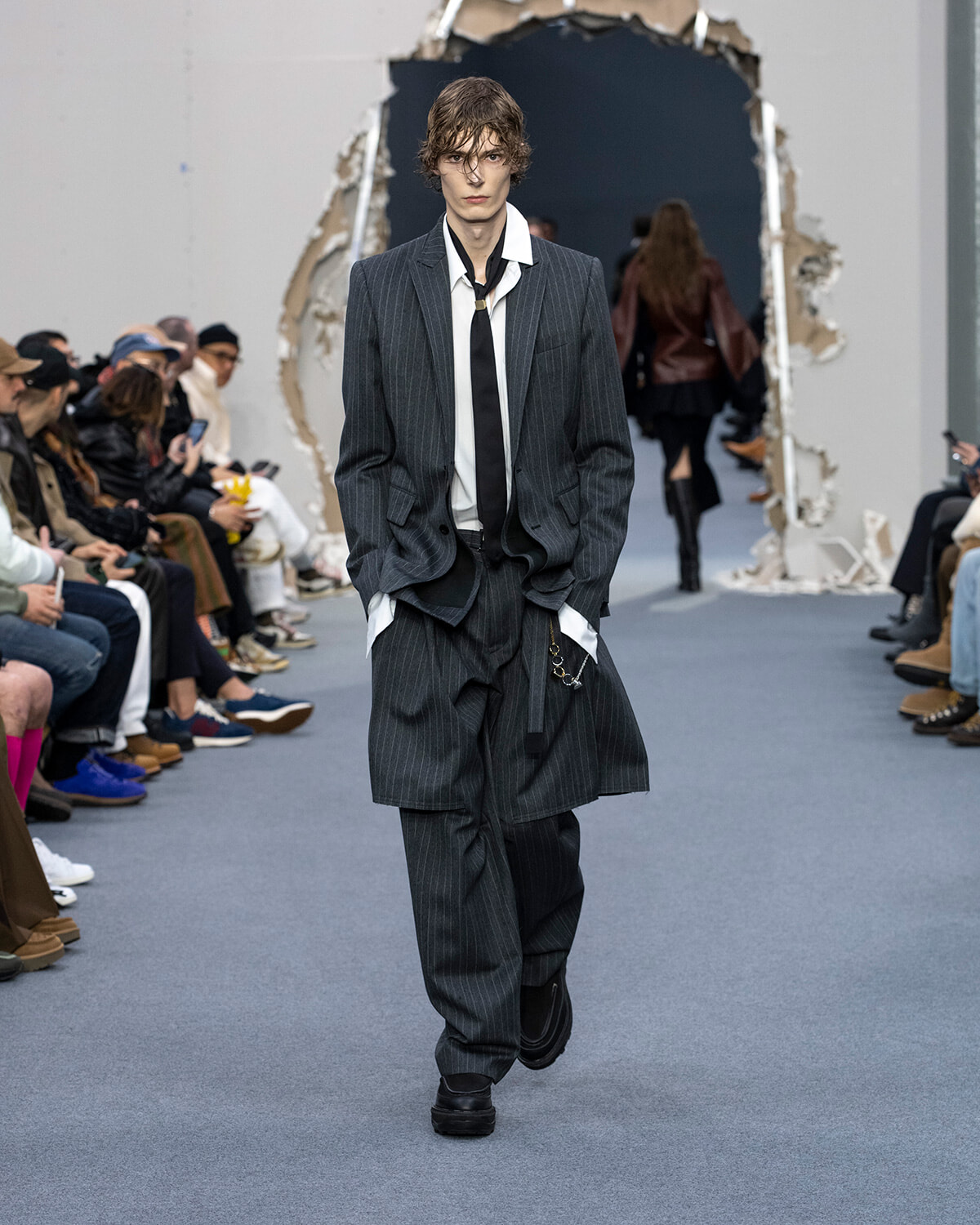 Male model walking a runway in a dark pinstripe suit with an oversized coat, white shirt, and black tie, hands in pockets as he strides forward.