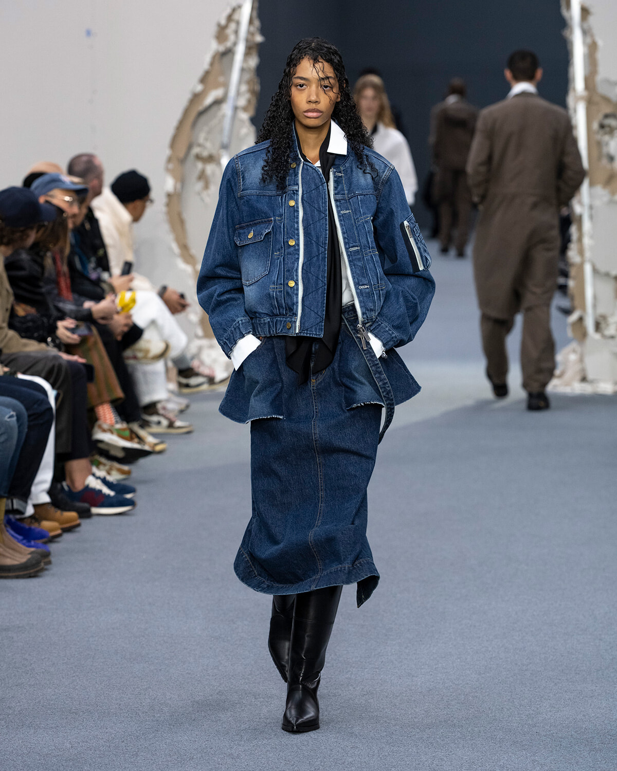 Model walks the runway in a double-denim outfit: oversized jean jacket over a long denim skirt, black boots, and layered white shirt accents.