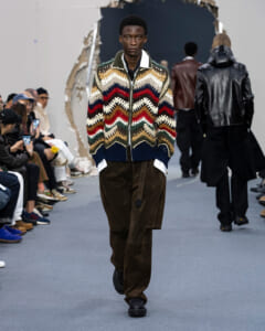 Male model walks a runway in a multicolor zigzag knit cardigan and brown cargo pants during a fashion show. Audience seated along the sides.