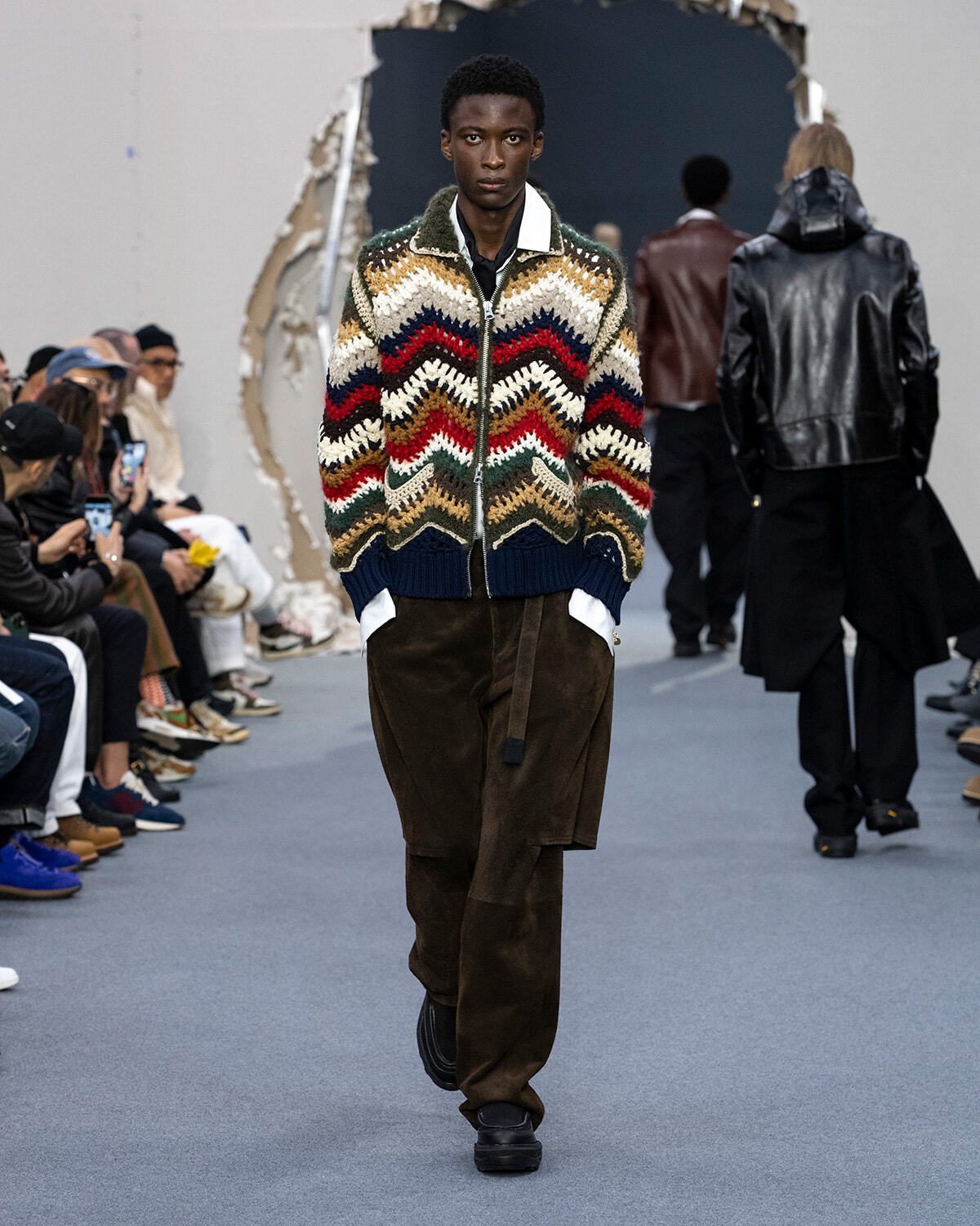 Male model walks a runway in a multicolor zigzag knit cardigan and brown cargo pants during a fashion show. Audience seated along the sides.