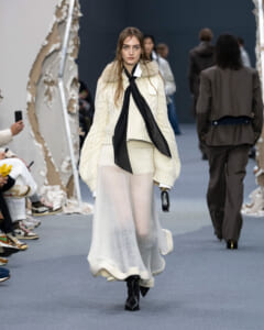 Model walks a fashion show wearing an ivory oversized knit sweater with a long black scarf and a sheer white skirt, black boots, and a small black clutch.