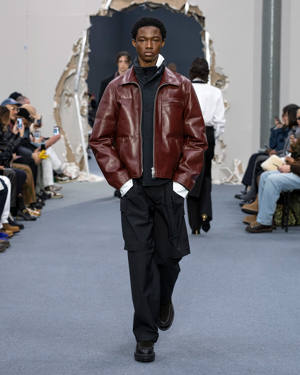 Male model on a runway wearing a burgundy leather jacket over a black zip-up and black cargo pants during a fashion show.