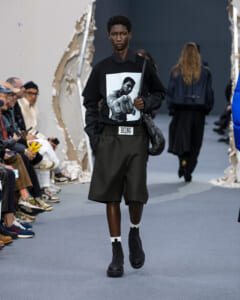 Male model walks a runway in an all-black outfit: graphic black T-shirt, black shorts, white socks, black boots, crossbody bag, and a BEING belt buckle.