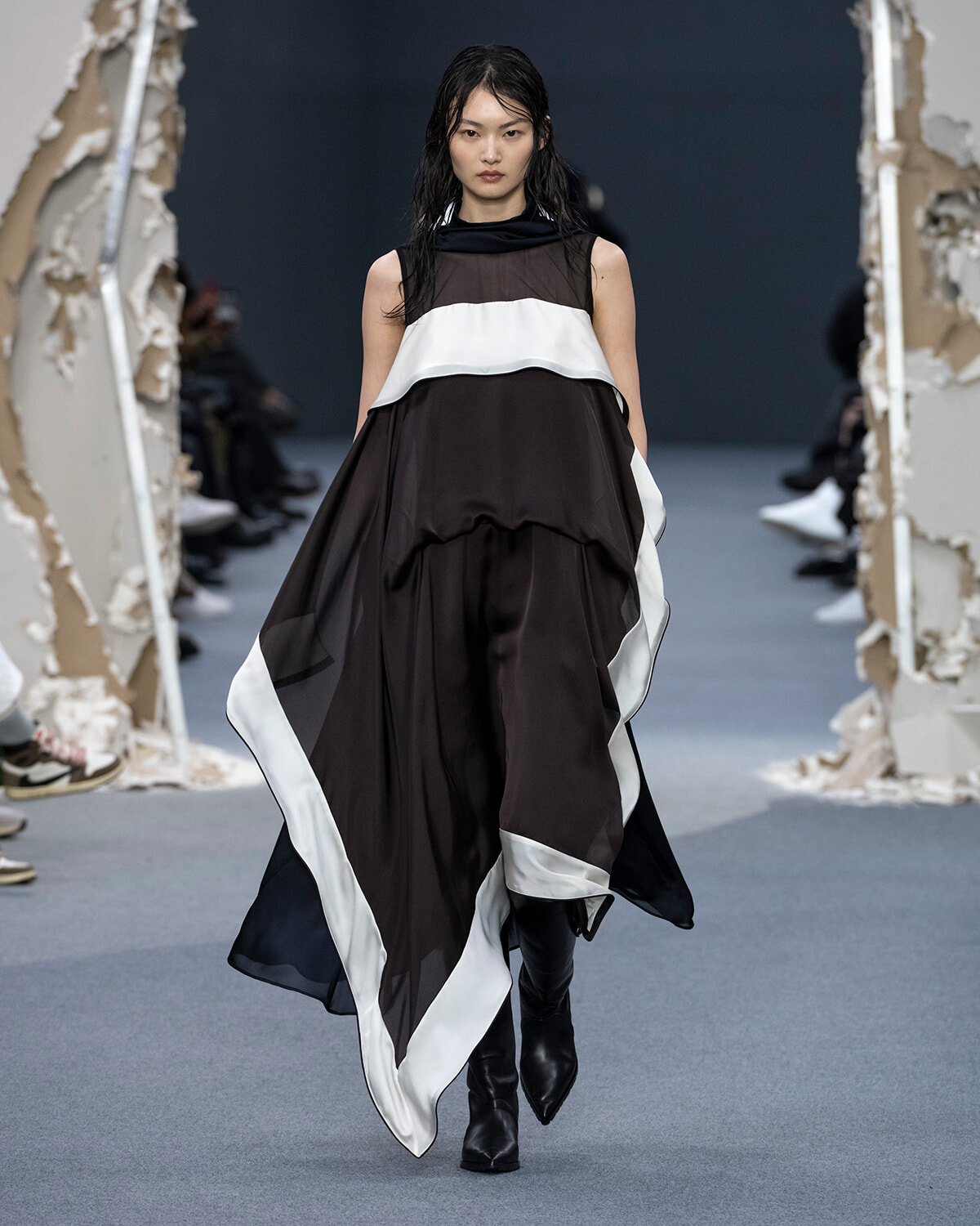 Model walks a fashion show wearing a dramatic black and white layered gown with sheer top and knee-high boots.