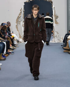 Model walks the runway in a dark brown shearling coat and matching pants with teal zipper accents, audience on both sides.