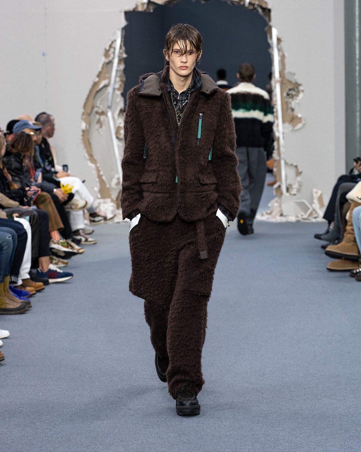 Model walks the runway in a dark brown shearling coat and matching pants with teal zipper accents, audience on both sides.