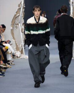 Model walks the runway wearing a color-block striped cardigan over a white shirt, paired with loose gray trousers and black shoes.