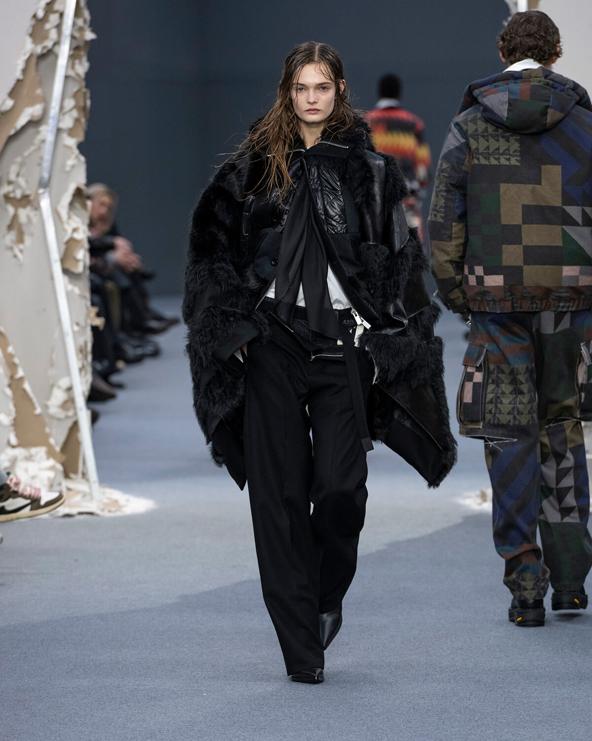 Female model walks the runway in an oversized black fur coat over a black suit, with a scarf.