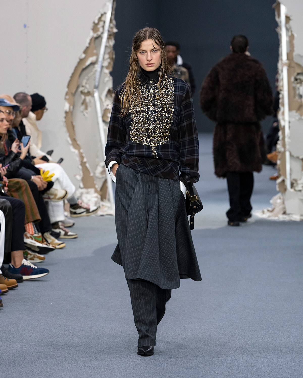 Model walks a fashion runway in a dark plaid top with dense bead embellishment over a layered pinstripe skirt; audience sits along the sides reading or watching.