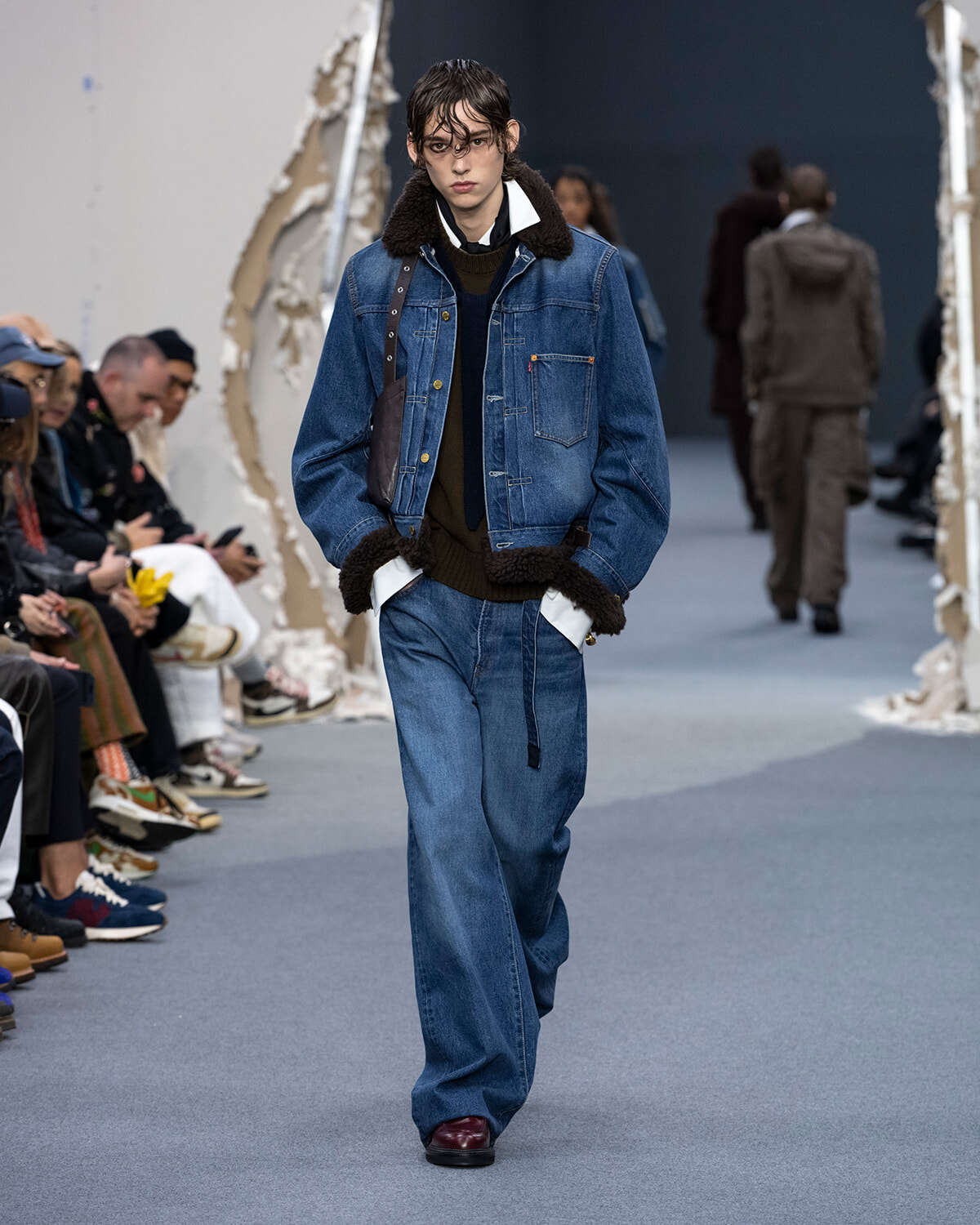 Model walking a fashion runway in an oversized denim jacket with shearling trim, layered with a dark sweater and wide jeans.