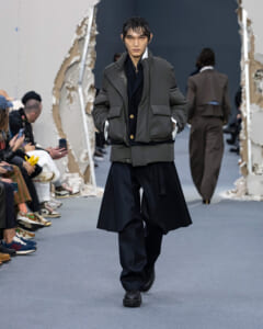 Male model on a fashion runway wearing a dark gray quilted puffer jacket over a navy coat and wide-leg navy pants; audience seated along the left side.