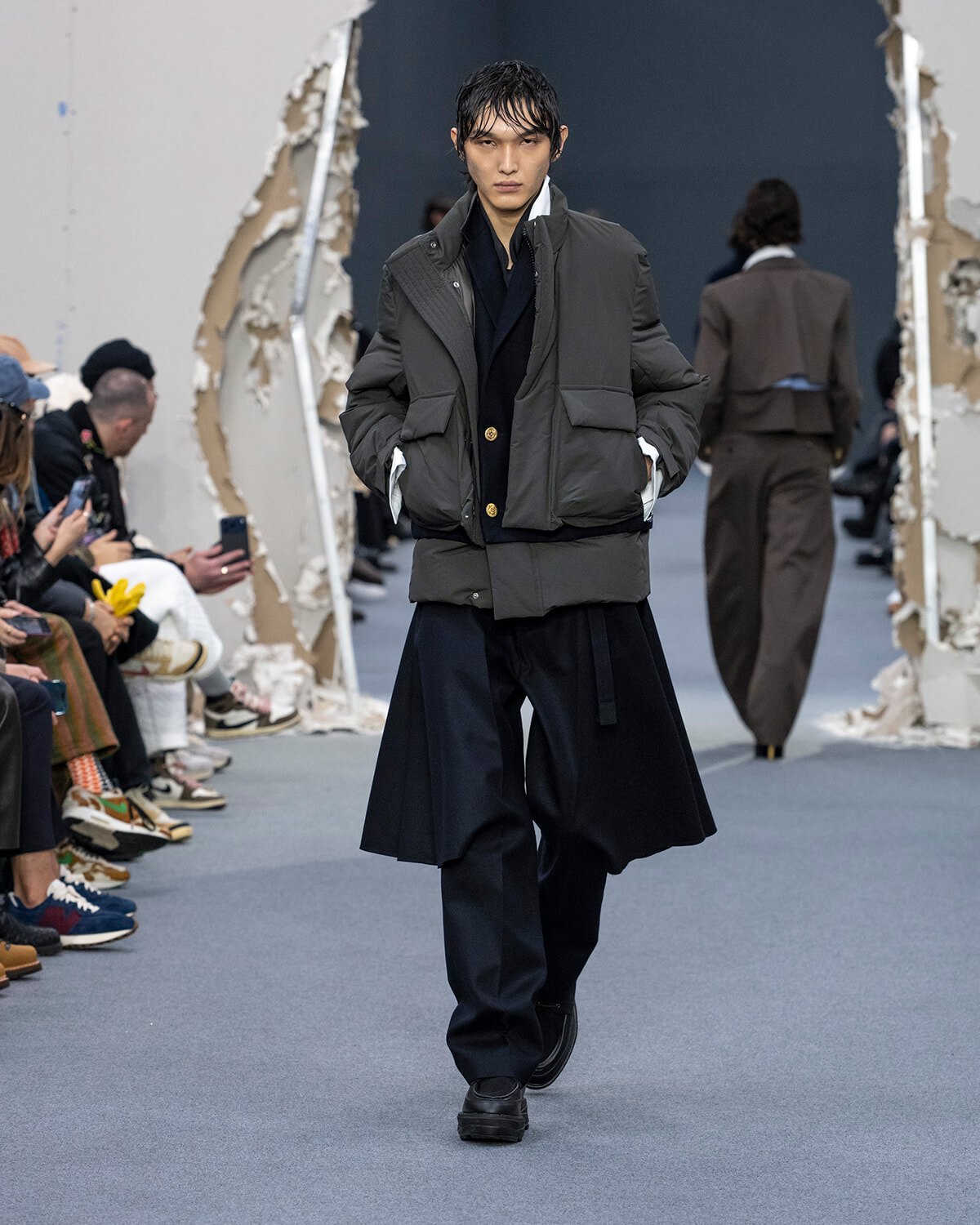 Male model on a fashion runway wearing a dark gray quilted puffer jacket over a navy coat and wide-leg navy pants; audience seated along the left side.