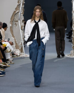 Model walks a runway in an oversized white shirt with a black neck-tie scarf and wide blue jeans, hands in pockets.