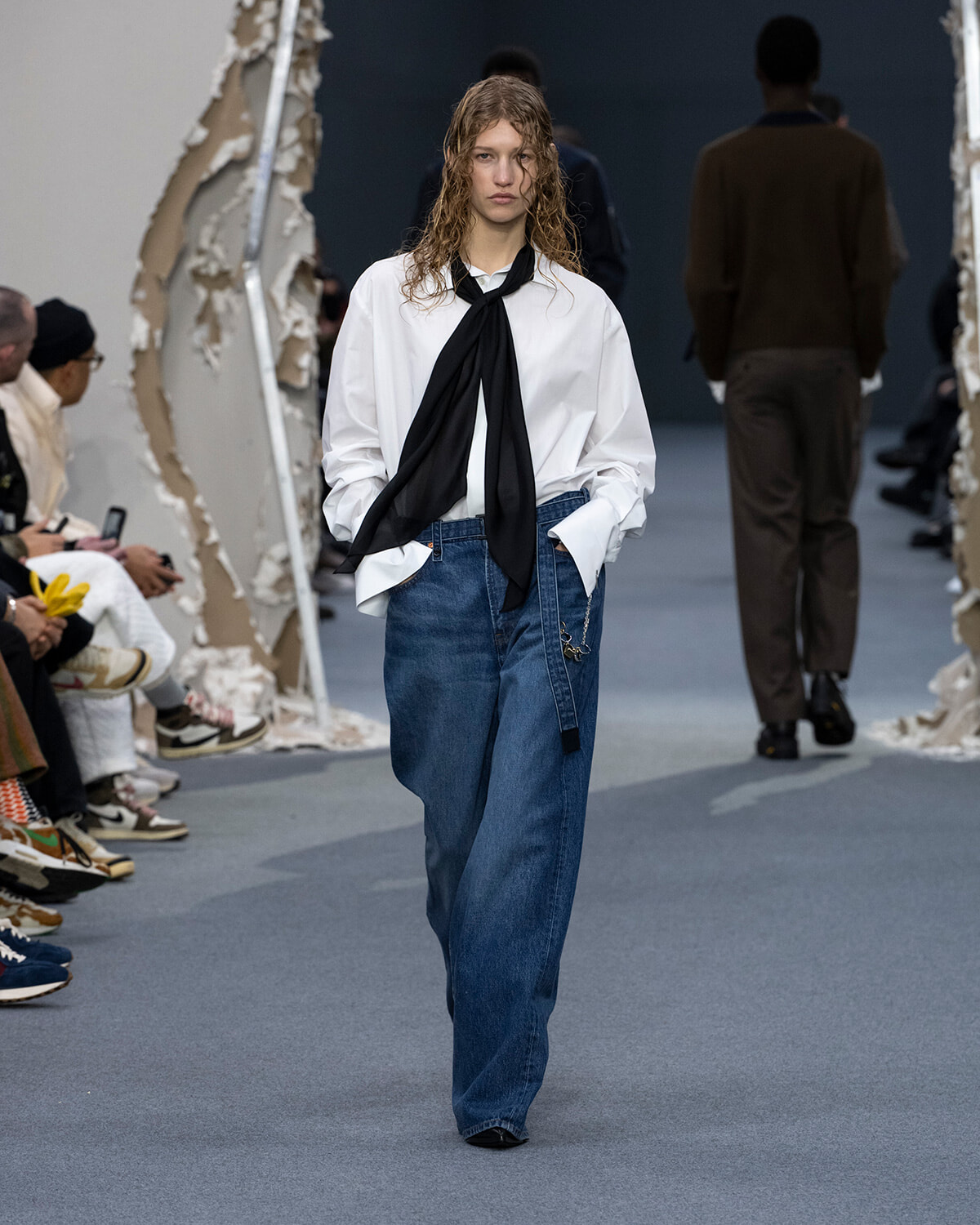 Model walks a runway in an oversized white shirt with a black neck-tie scarf and wide blue jeans, hands in pockets.