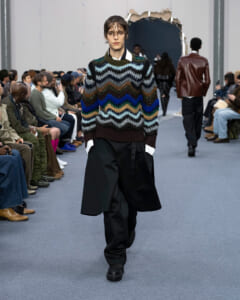 Model walks the runway in a chunky multicolor chevron knit sweater and oversized black trousers as the audience looks on from the sides.