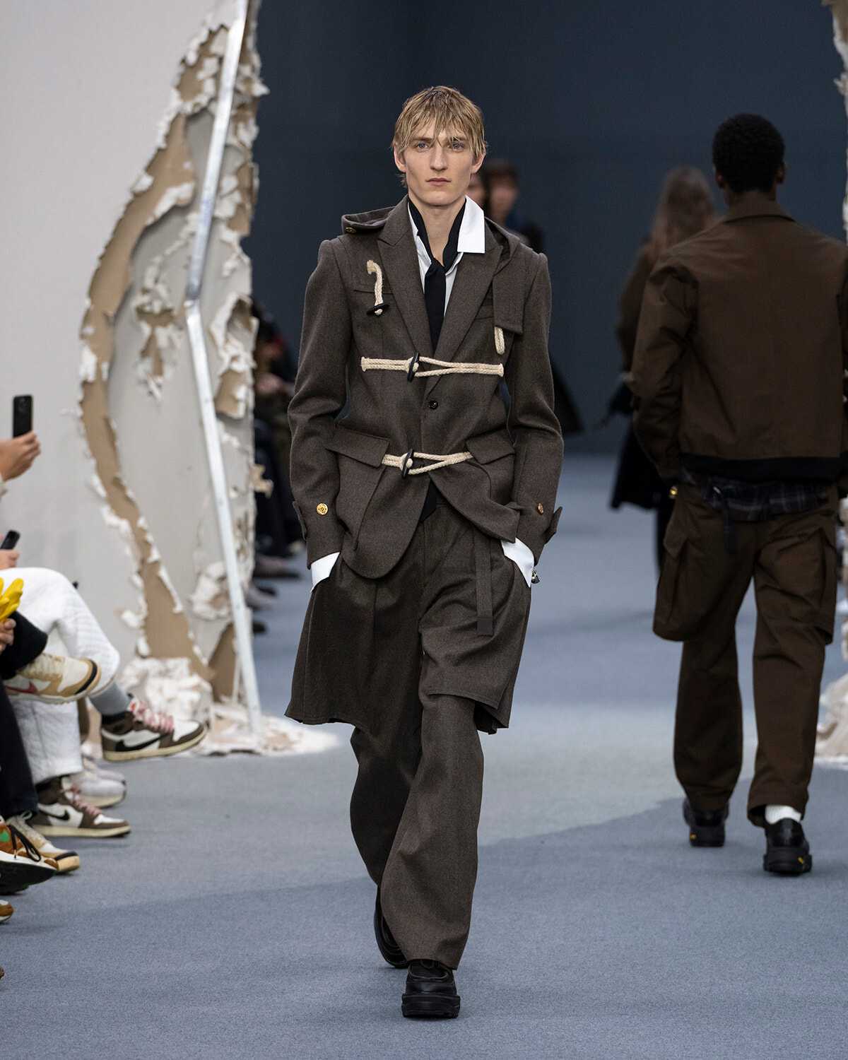 Model walks the runway in a brown layered suit with rope belts and a white shirt, during a fashion show.