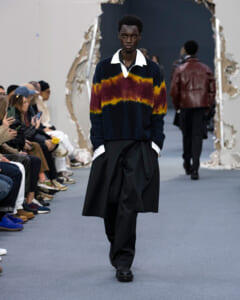 Male model on a runway wearing a chunky knit navy sweater with yellow and rust color bands, white collar peeking out, and oversized black pants; audience seated along the catwalk.Background shows a large torn-wall backdrop.