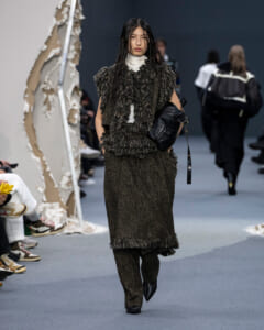 Model walks the runway in a dark brown, fringed knit set with a white turtleneck, carrying a black bag, on a gray catwalk.