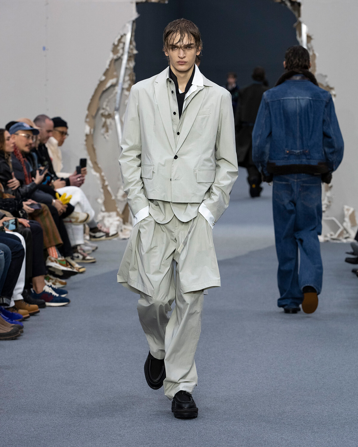 Male model in a pale gray layered suit walks the runway, hands in pockets, spectators seated along the sides.