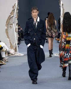 Male model walks the runway in a navy denim suit with white shirt and black tie, hands in pockets.