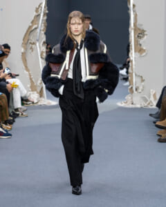 Model walks the runway in an oversized black-and-white fur jacket over black layers, with a serious expression.