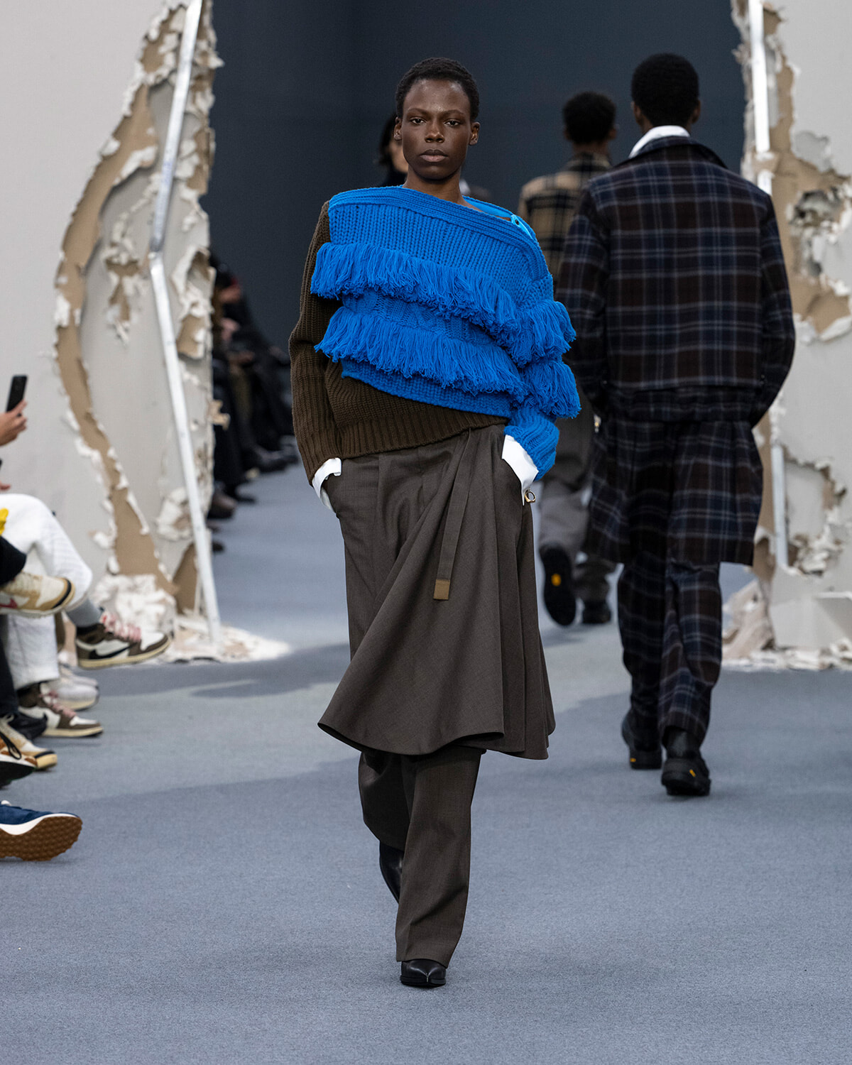 Model walking a fashion runway in a bright blue fringe-knit sweater layered over brown knitwear and wide brown pants, with audience seated along the sides.