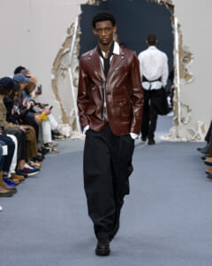 Male model walks a runway wearing a brown leather blazer, white shirt, black wide-leg trousers, and black shoes.