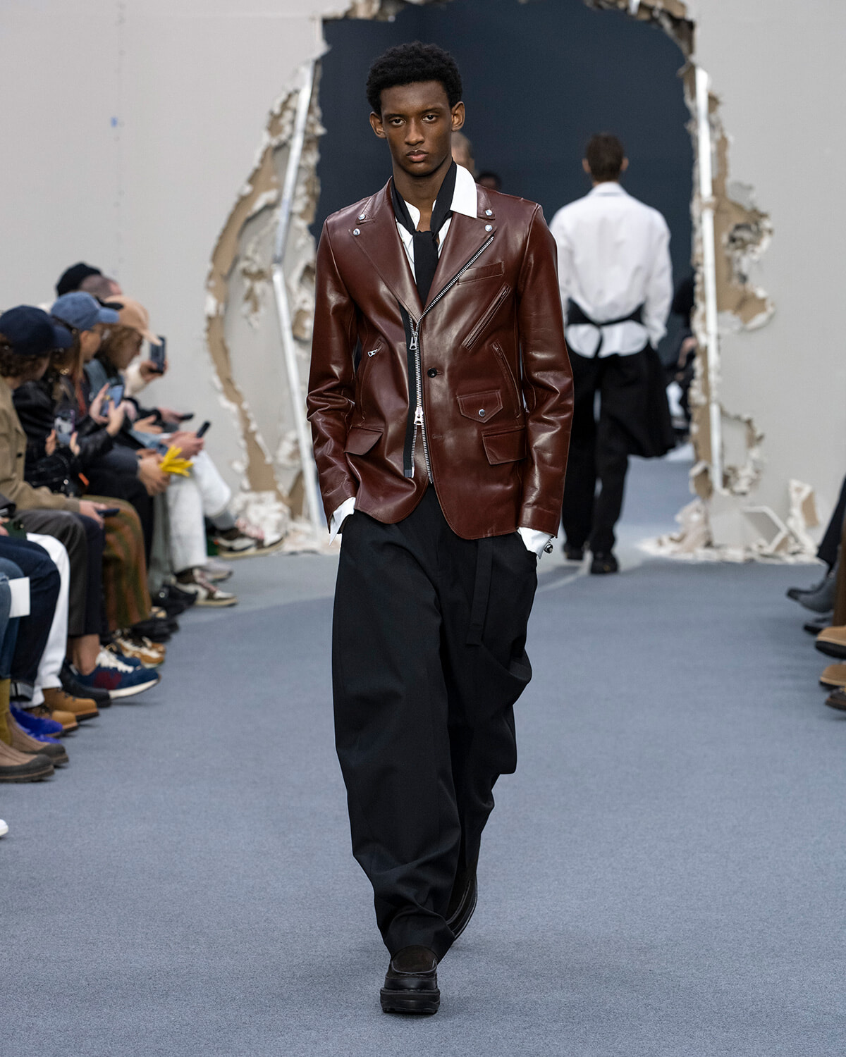 Male model walks a runway wearing a brown leather blazer, white shirt, black wide-leg trousers, and black shoes.