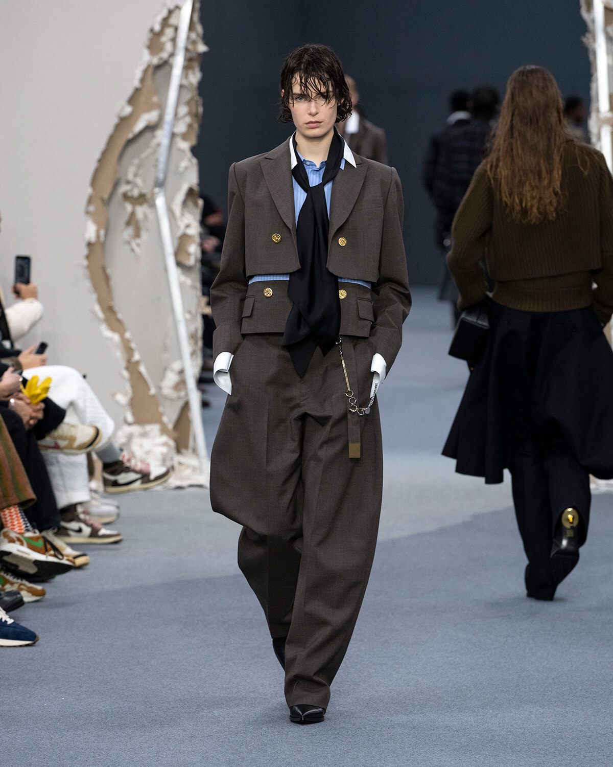 Model walking the runway in an oversized brown suit with gold buttons, blue shirt and black scarf.