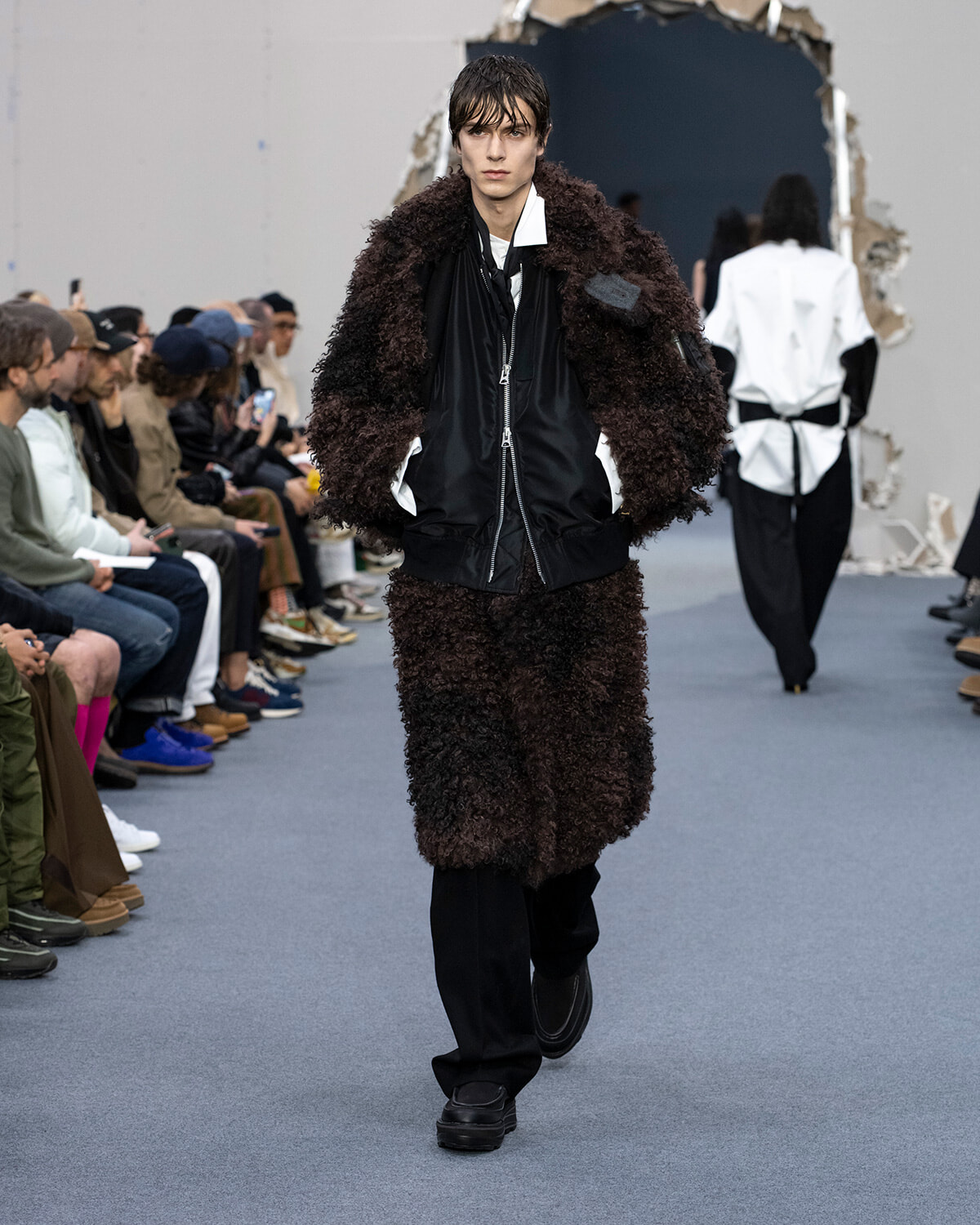 Model walking a fashion show in a brown shaggy coat over a black zip jacket and black pants, with spectators on both sides of the runway.