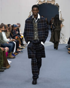 Male model walking a runway in a dark plaid suit with matching jacket and wide pants, white shirt, and a patterned vest. Audience on both sides.