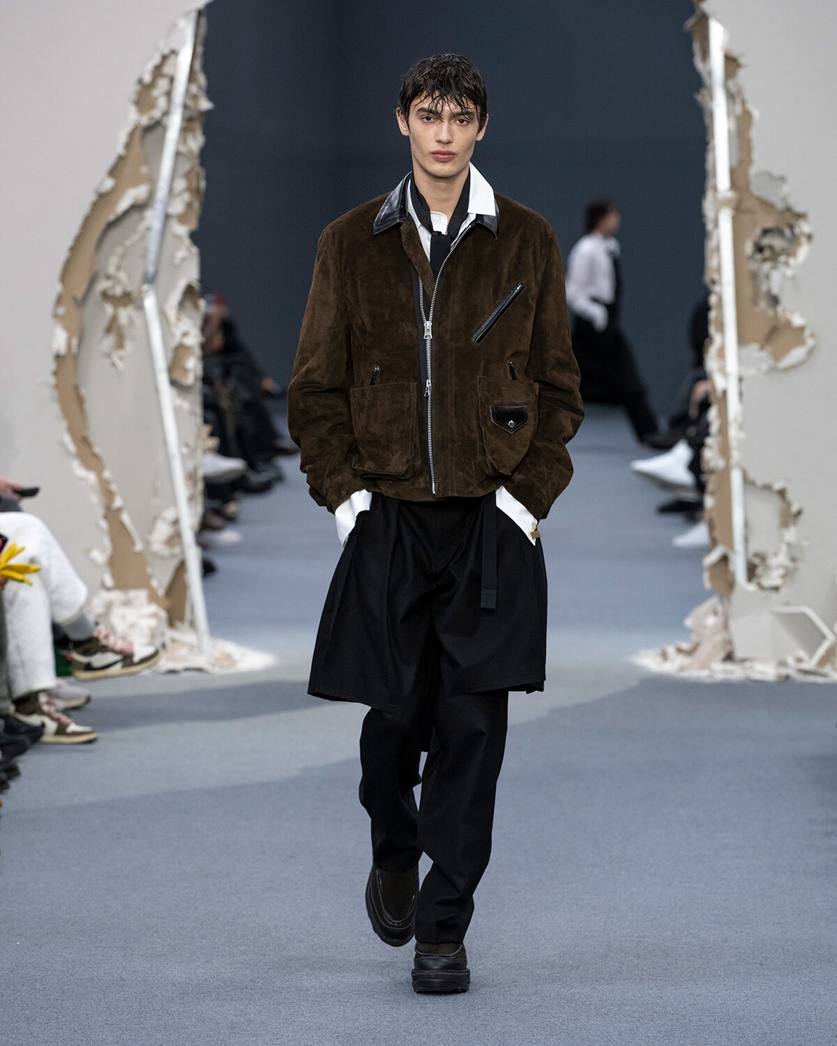 Male model walks a runway wearing a brown suede zip-front jacket with multiple pockets over a white shirt and black layered trousers.