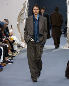 Model walking the runway in a brown suit with layered blue denim shirt/jacket, hands in pockets, audience on both sides.