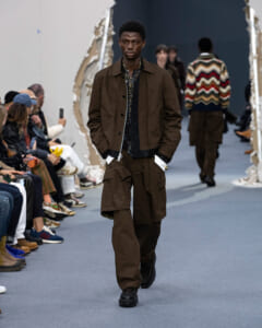 Male model walks the runway in a brown utility jacket and cargo pants, audience on both sides of the catwalk.