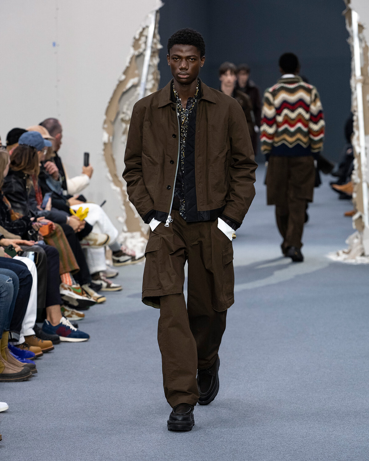 Male model walks the runway in a brown utility jacket and cargo pants, audience on both sides of the catwalk.