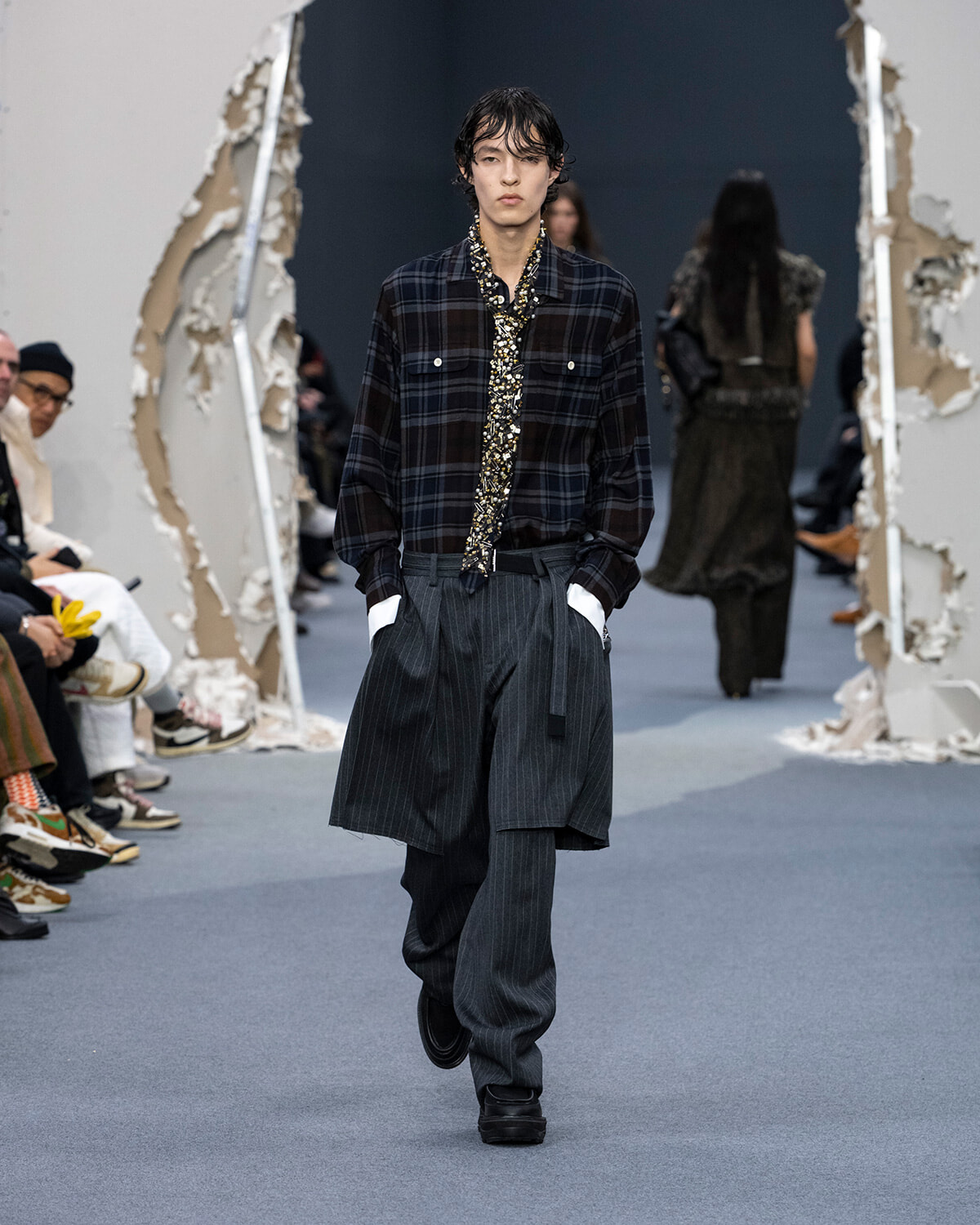 Model walks down a fashion runway in an oversized dark plaid shirt with a floral scarf, wide pinstriped layers, and black shoes, audience on sides.