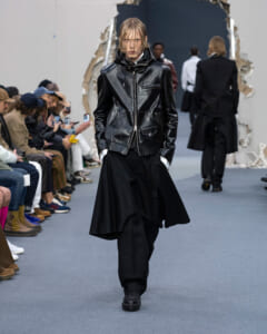 Model walks the runway in a black leather zip-front jacket with large pockets, paired with oversized black pants, audience at left watching.