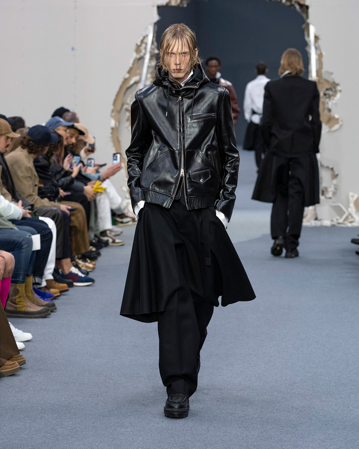 Model walks the runway in a black leather zip-front jacket with large pockets, paired with oversized black pants, audience at left watching.