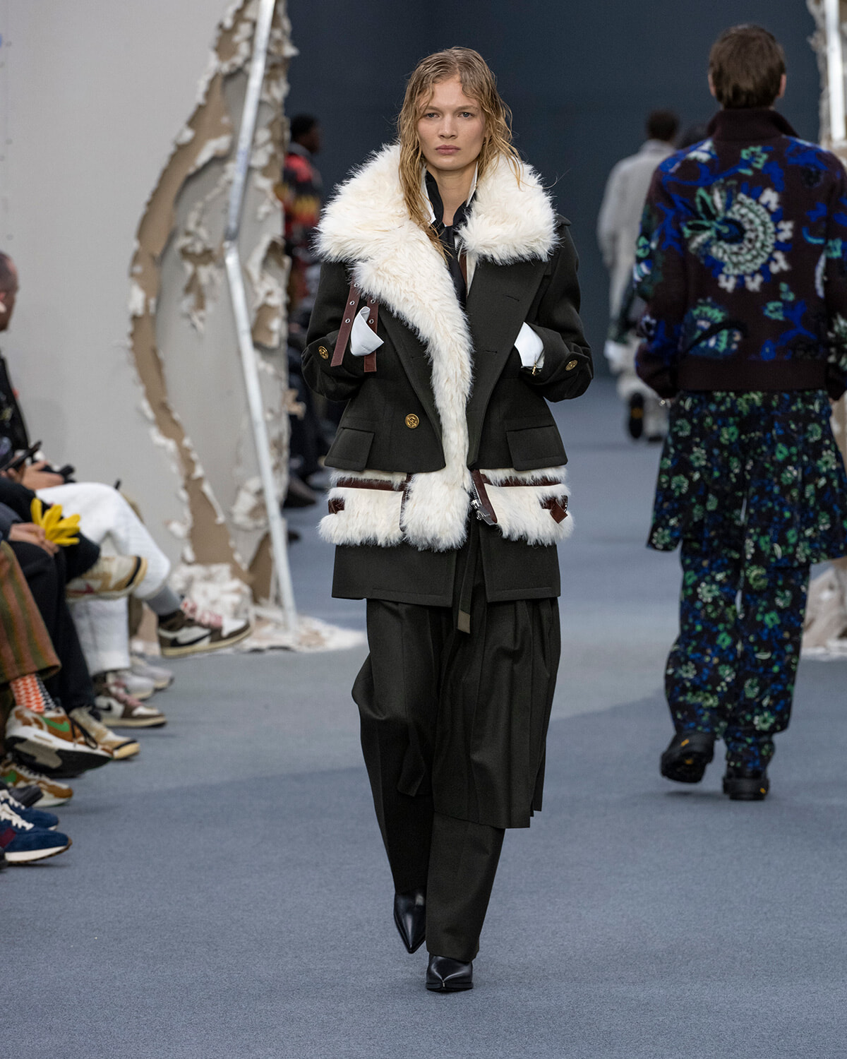 Model walking a runway in a black double-breasted coat with white faux-fur collar and hem, paired with wide-leg black pants and black boots.