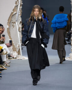 Model walking a fashion runway in a dark denim jacket, white shirt, black scarf, and wide black trousers with fur cuffs.