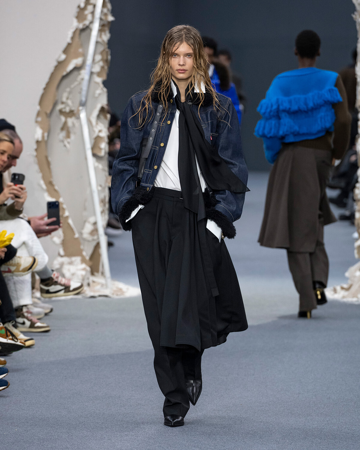 Model walking a fashion runway in a dark denim jacket, white shirt, black scarf, and wide black trousers with fur cuffs.