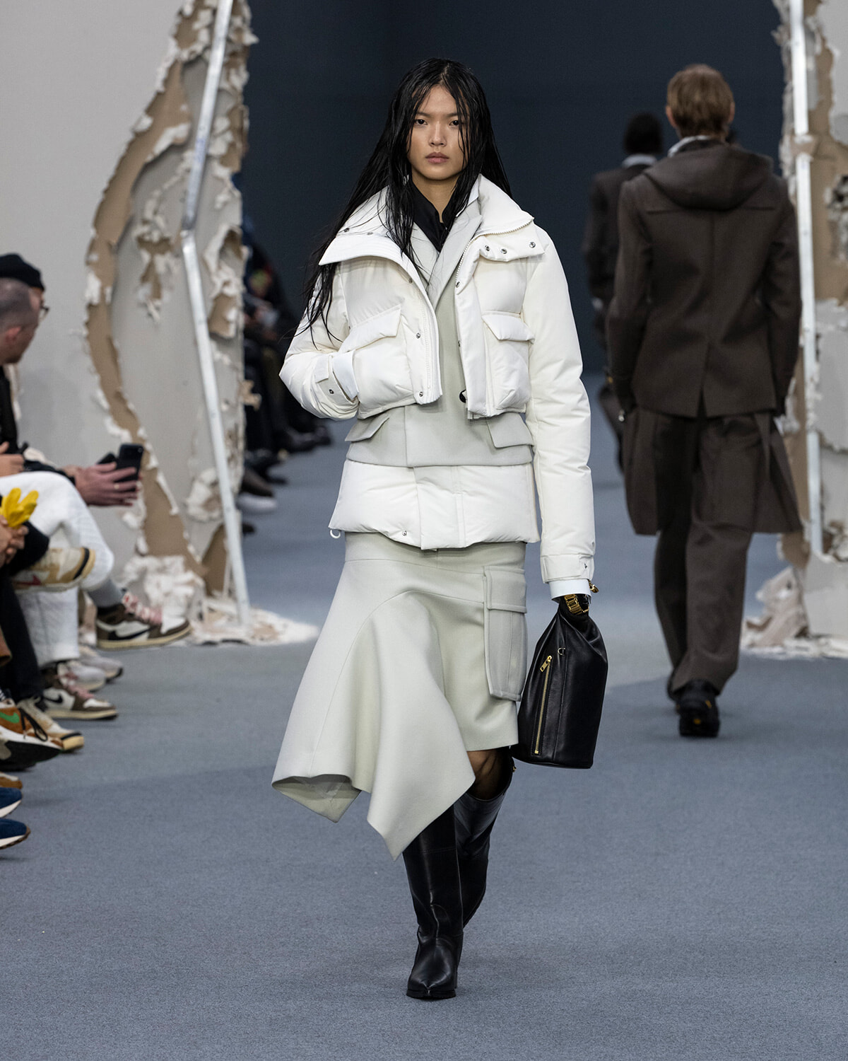 Model walks a runway in an oversized white puffer jacket, light gray layered skirt, and black knee-high boots, carrying a black handbag.