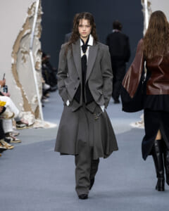 Model on a runway wearing a grey tailored suit with a long layered overcoat, white shirt, and black necktie.
