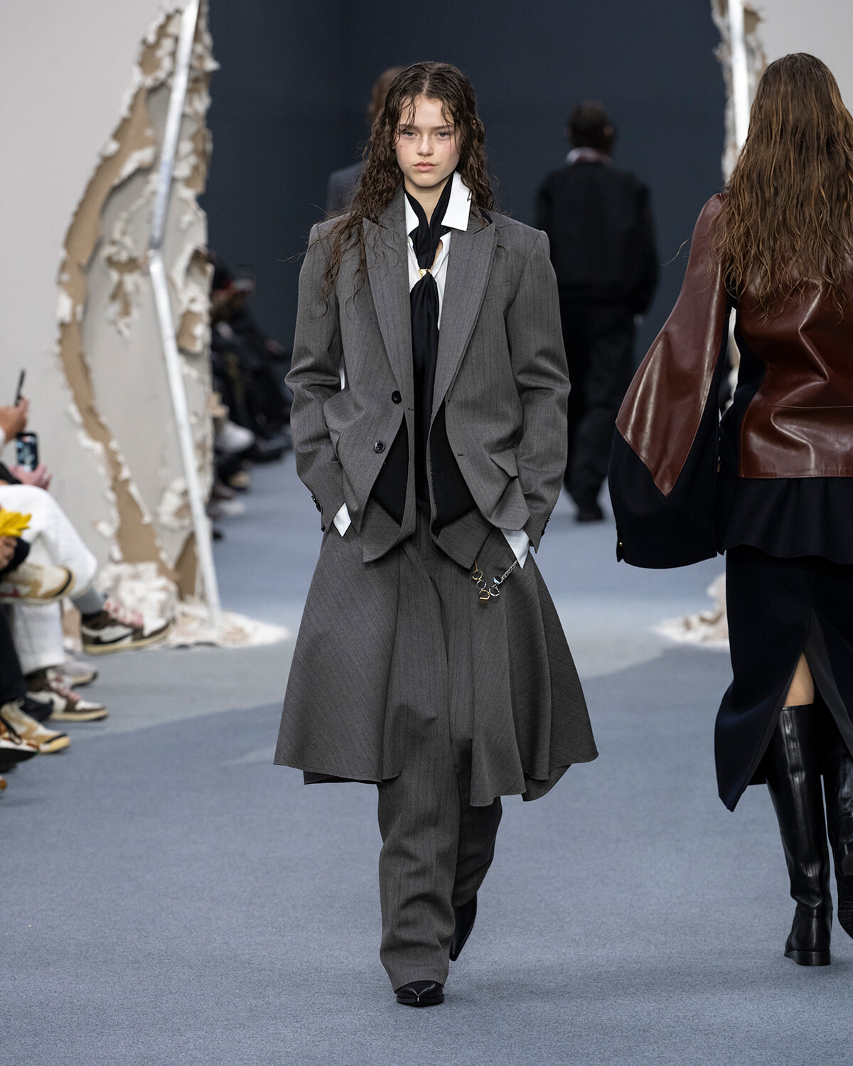 Model on a runway wearing a grey tailored suit with a long layered overcoat, white shirt, and black necktie.