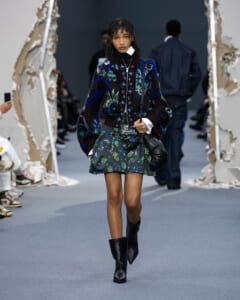 Female model walking a runway in a dark blue floral knit jacket, matching floral skirt, knee-high black boots, and a black bag across her body.