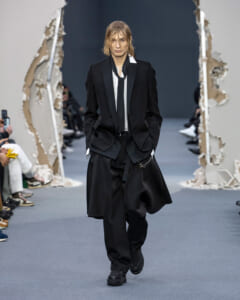 Model in an oversized black suit with a white shirt and black tie walks the runway with hands in pockets.